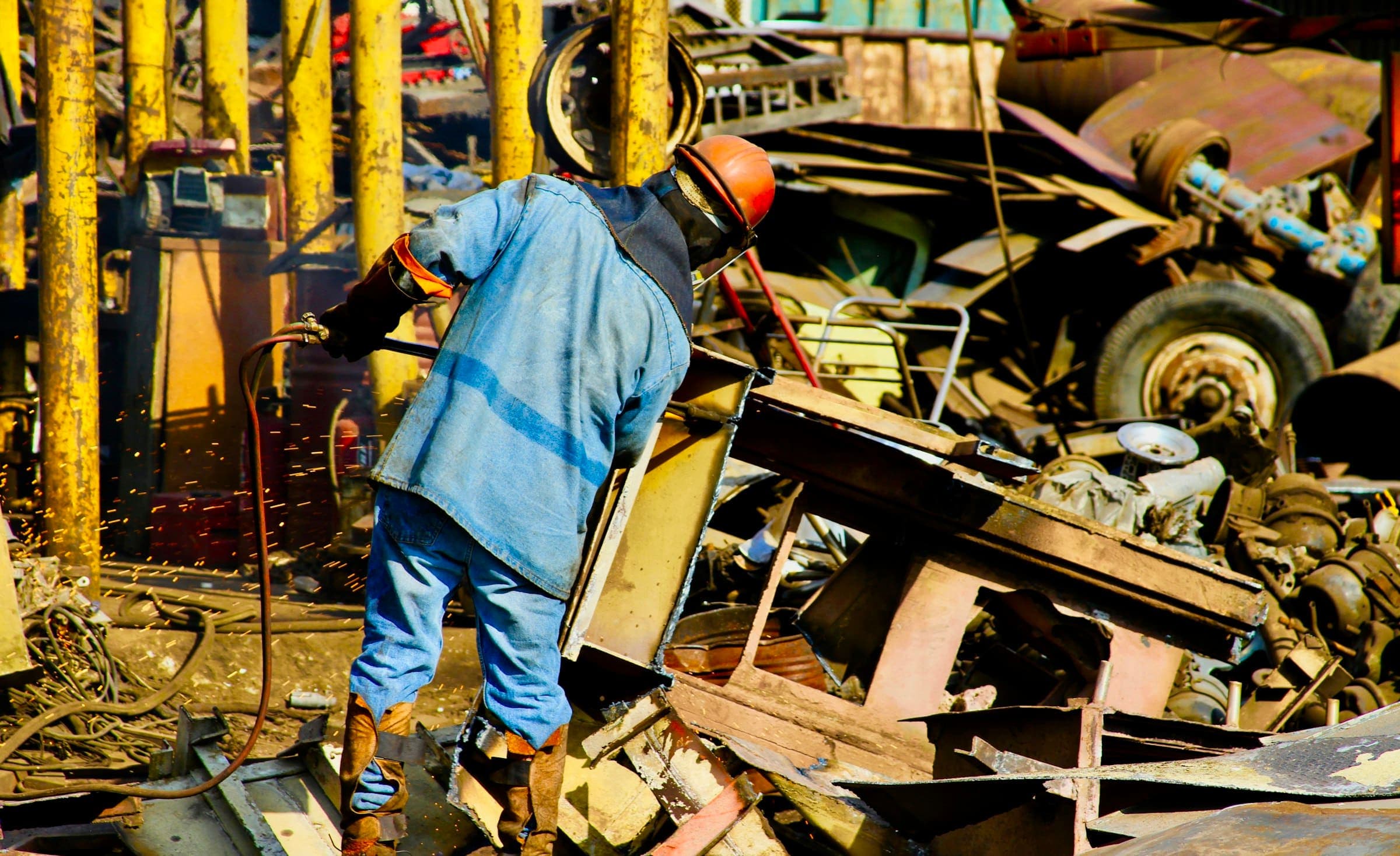 A worker in a hard hat cutting up scrap metal with a gas torch as sparks fly
