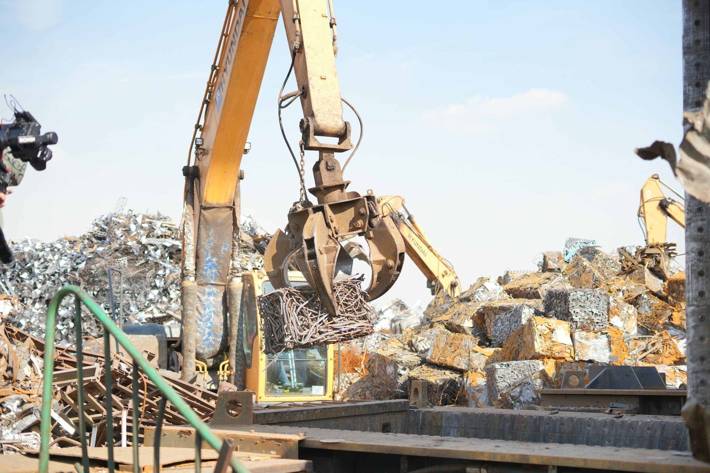 An excavator's grapple lifting a bundle of scrap metal in a scrap yard