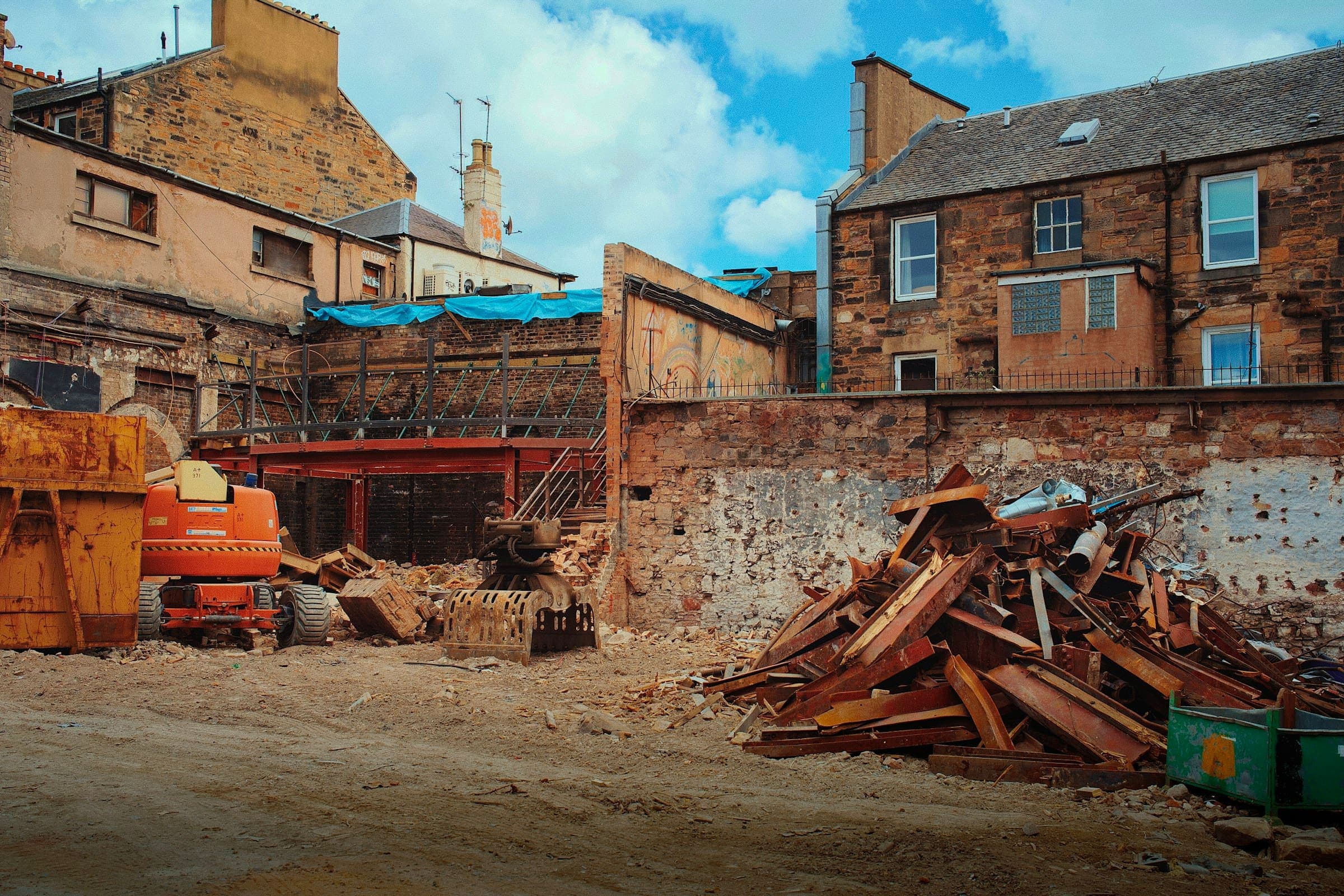 A heap of rusty steel beams on a cleared demolition site, with skips and a boom lift nearby