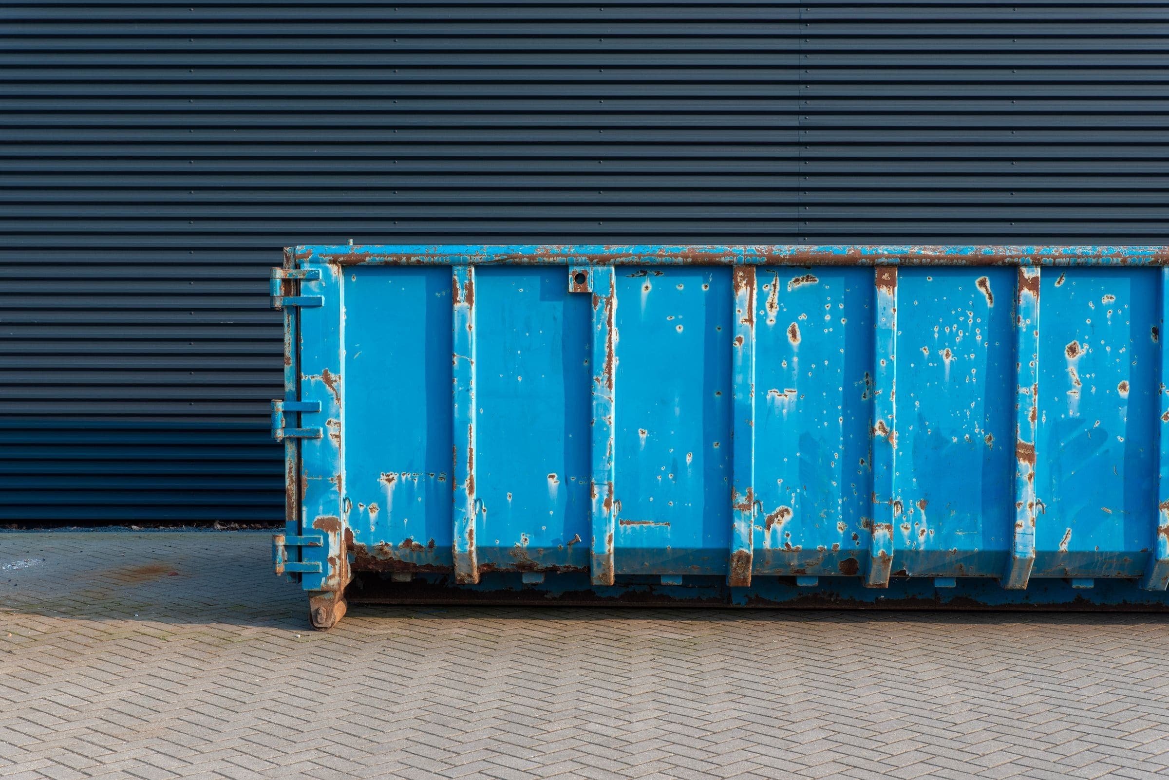 A long, rusty blue roll-off bin on brick paving in front of a dark corrugated metal wall A long, rusty blue roll-off bin on brick paving in front of a dark corrugated metal wall