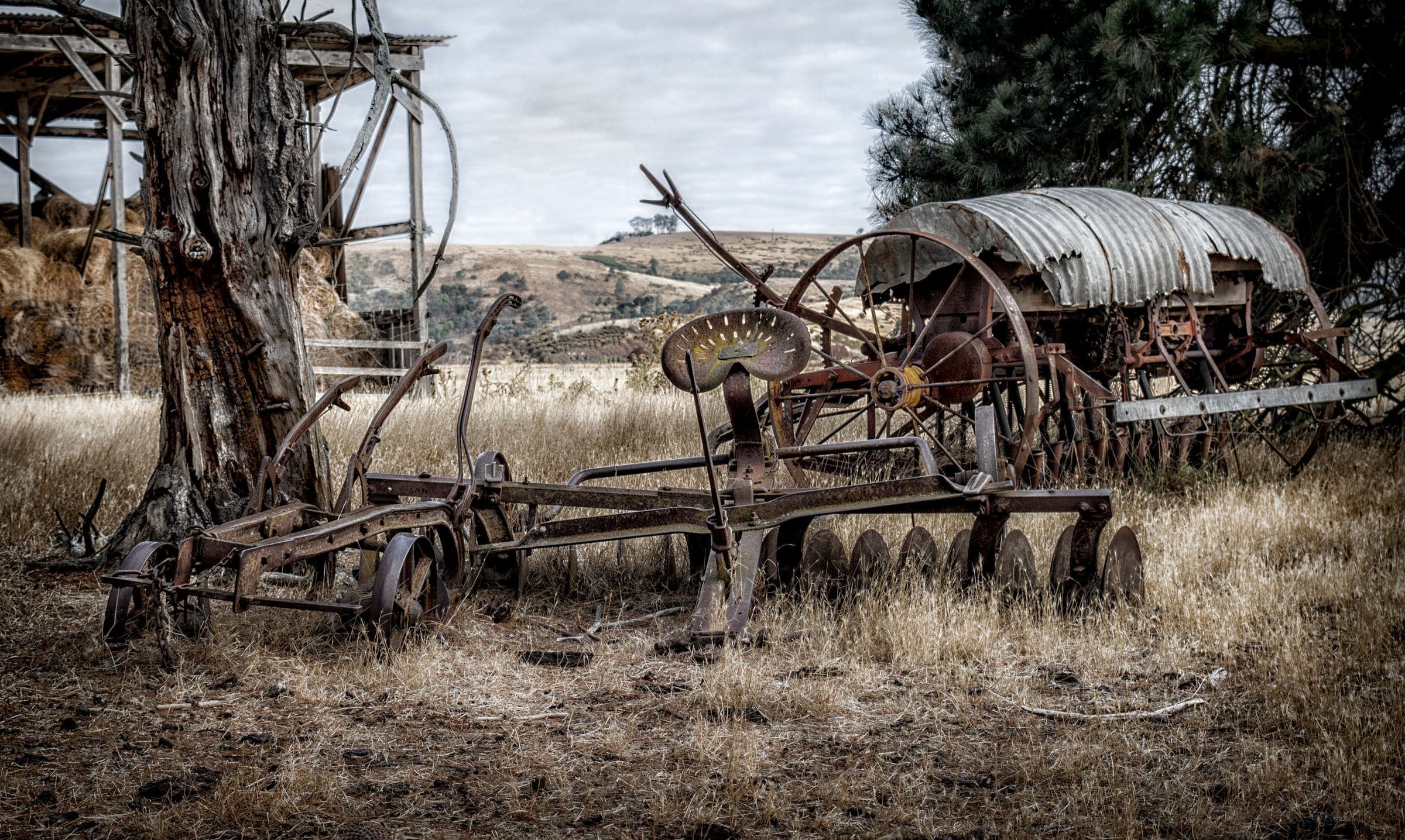 Old farm machinery Rusty old farm implements, including a disc plough and a seeder with an iron seat, in a dry paddock beside an old gum tree