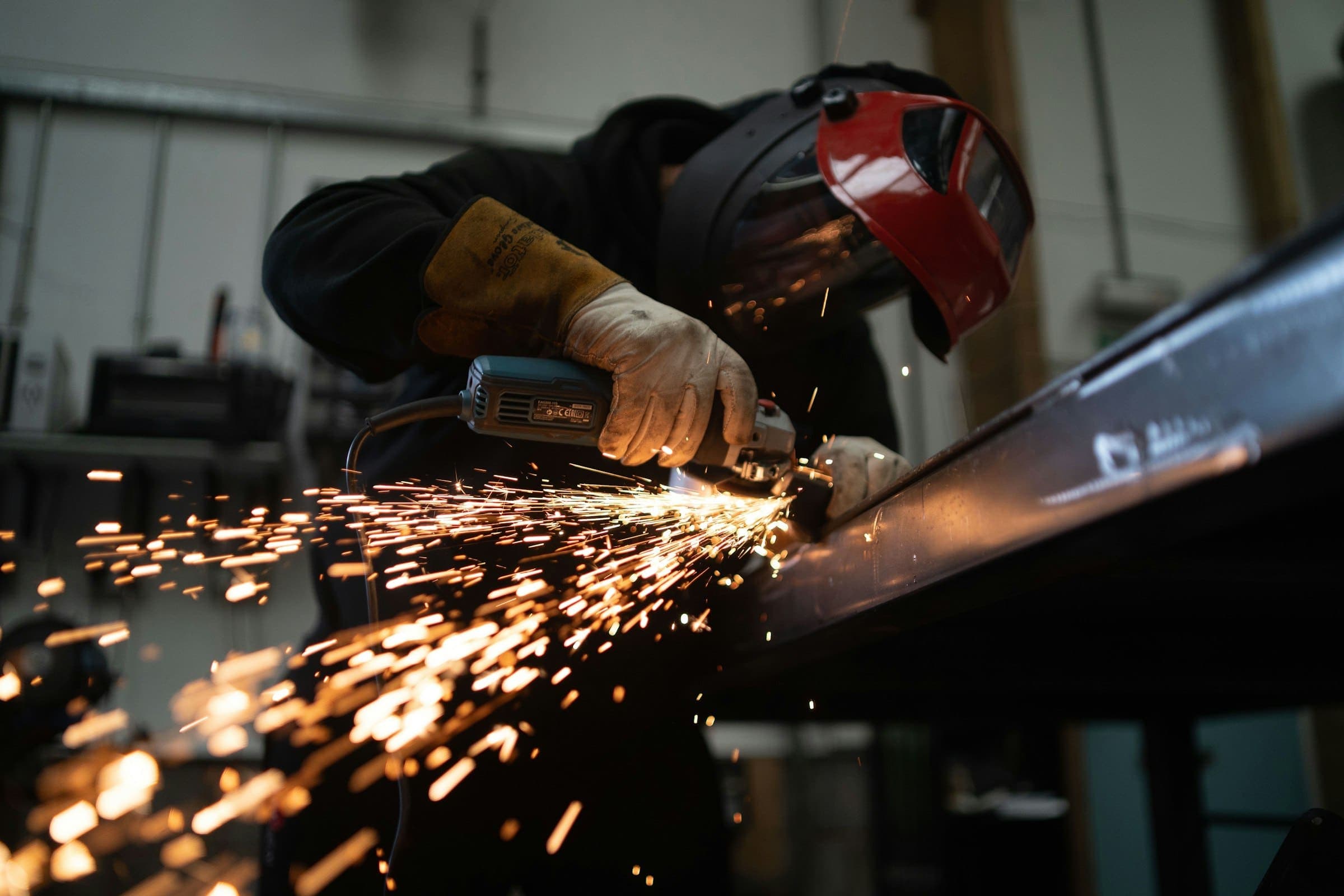 A worker in a welding helmet and leather gloves cuts a steel beam with an angle grinder, sending out a spray of sparks A worker in a welding helmet and leather gloves cuts a steel beam with an angle grinder, sending out a spray of sparks