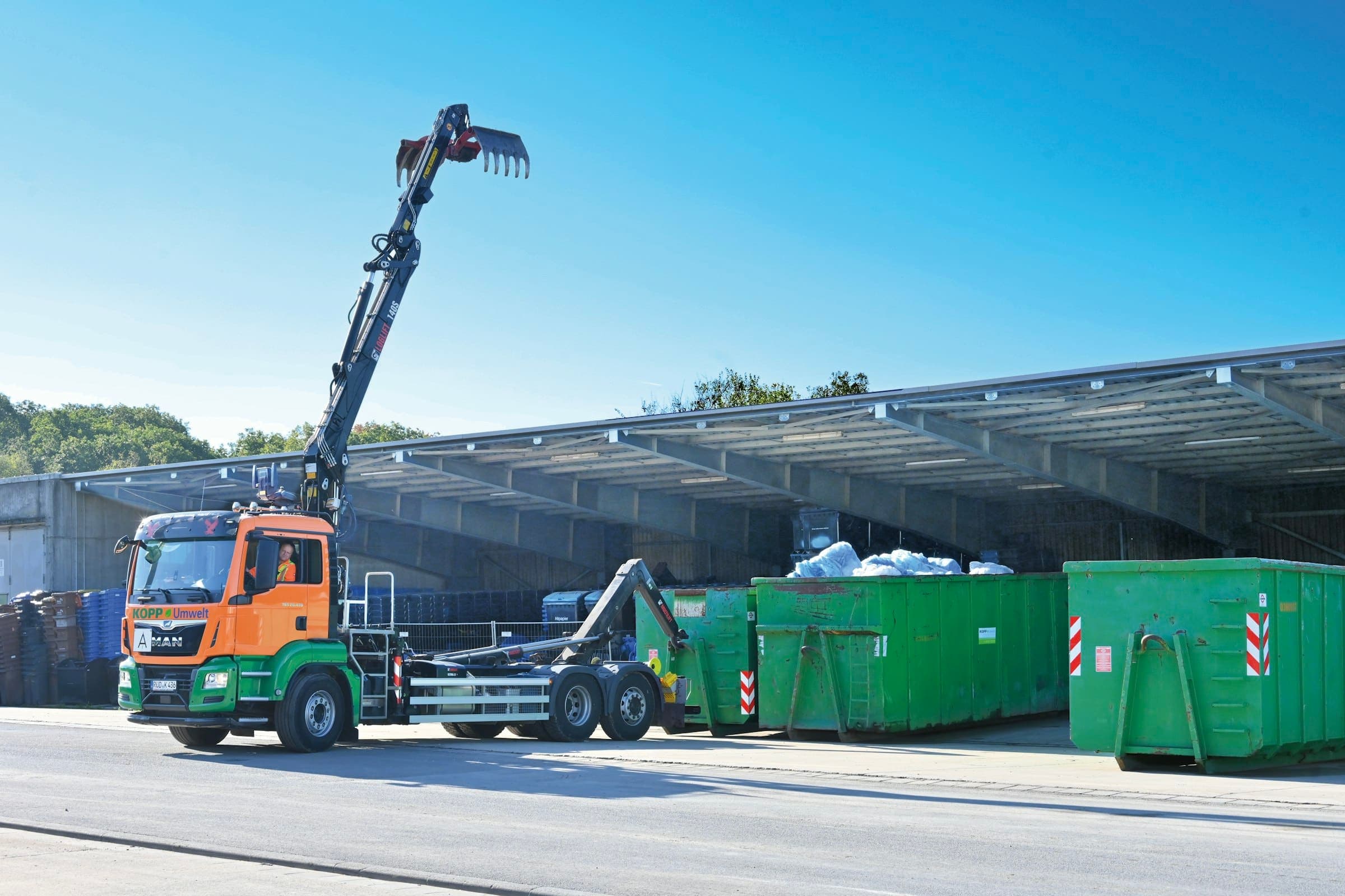 Hook-lift truck and bins A hook-lift truck with its crane raised beside a row of green roll-off bins outside a recycling shed